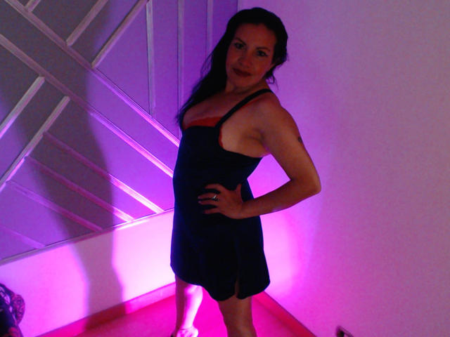 SashaBlue69 stands in a dark blue dress with orange trim, hands on hip, purple and pink lighting. She has long black hair, small tits, and a confident smile.