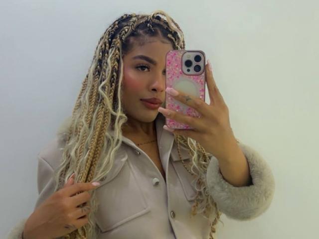 LexieBloom with long blonde braids, wearing a beige button-up shirt, holding a pink phone with white dots, pouting lips, cute face, and slightly flushed cheeks, showing off nails.