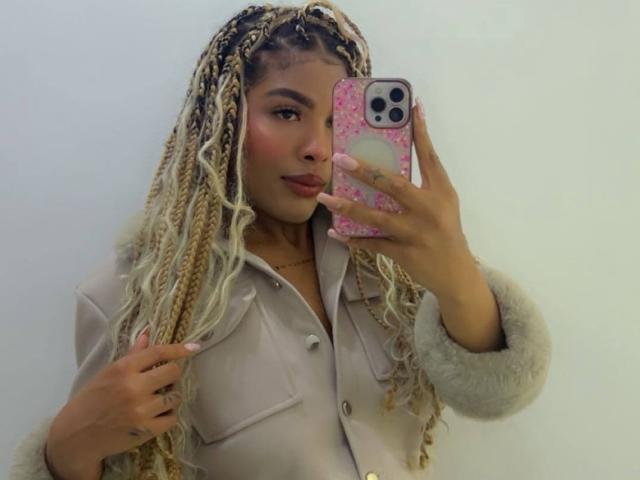 LexieBloom con trecce bionde, labbra carnose, custodia rosa per il telefono, giacca beige, che tiene il telefono, ammiccando, fard rosa, parete bianca, che si guarda allo specchio, mostrando il seno..