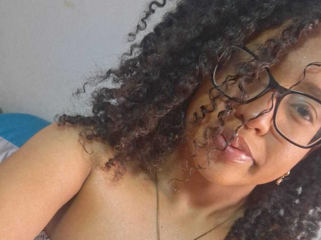 Artemisa, met krullend zwart haar, een bril op, haar tong uitstakend, zonder top, waarbij haar kleine tot middelgrote borsten zichtbaar zijn, bruine huid en een lichte glimlach, op een selfie..