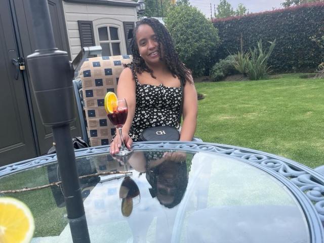 ArtemisaS con i capelli neri ricci, indossa una maglietta a pois bianchi e neri, tiene un bicchiere con una bevanda rossa e una fetta d'arancia, è seduta a un tavolo con piano in vetro e c'è un ombrello nero..