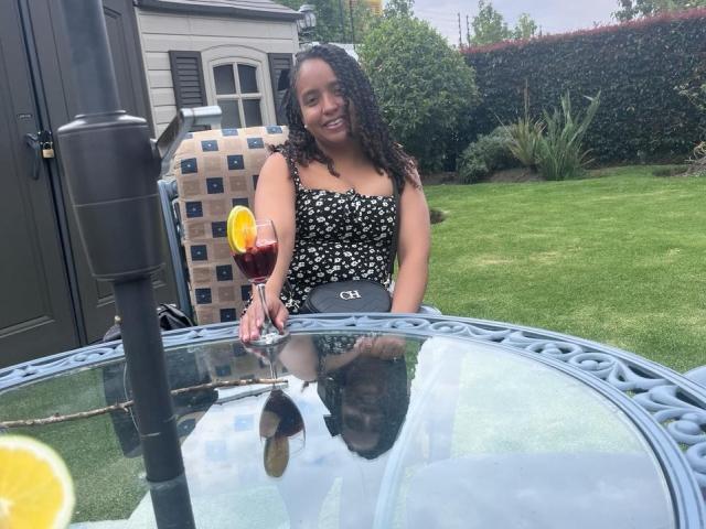Artemisa, met krullend zwart haar, draagt een zwart-wit gestreept jurk en zit aan een glazen tafel met een drankje. Haar reflectie is te zien in het glas, en de scène speelt zich buiten af..