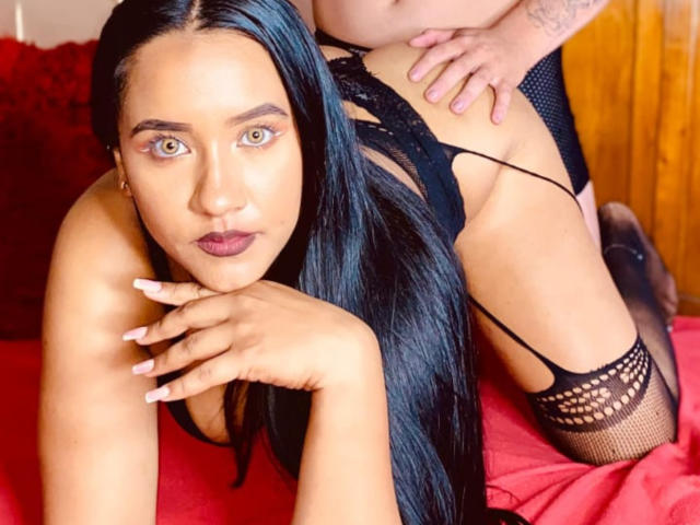 VelaAndEdwar with long black hair, brown eyes, and dark lipstick, wearing black lace lingerie, on red bed, looking at camera, with hands on bed, and another person's hand on ass.