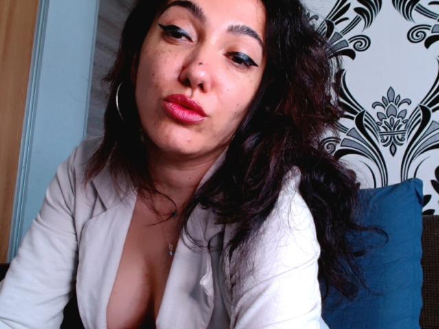 SexyMirra with dark curly hair, wearing a white shirt, pouting red lips, and black eyeliner, showing cleavage, sitting with a blue pillow. Her ass is slightly visible.