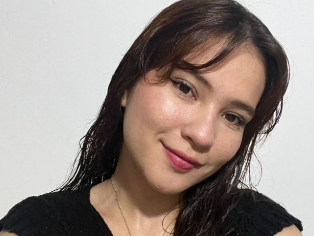 CelesteDash has wet brown hair, pink lipstick, and a slight smile, wearing a black shirt with a lace pattern, and a thin necklace, against a plain white background.