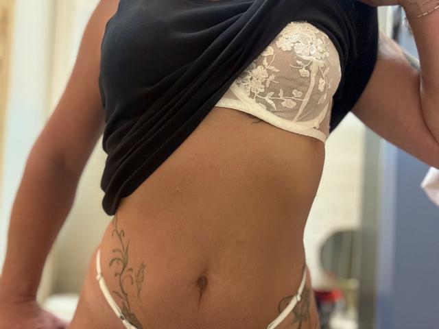 CoquineX69's torso, showing a black shirt lifted to reveal white lace bra, exposing a flat stomach with a navel and floral tattoos on the right side, wearing white panties.