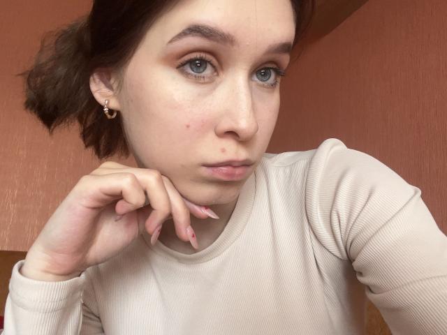 BellaBoom with pale skin, blue eyes, and brown hair in a ponytail, wearing a beige long-sleeve shirt, has a pensive expression, resting her chin on her hand.