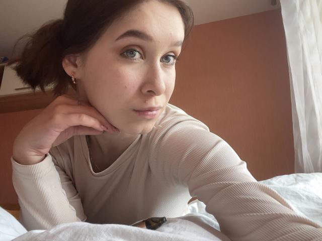 BellaBoom with blue eyes, light skin, and brown hair in a ponytail, wearing a beige ribbed shirt, resting her chin on her hand, looking at the camera.