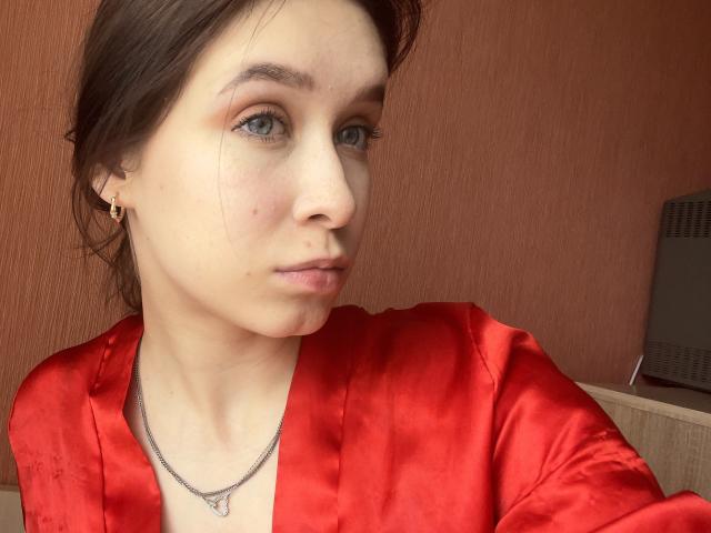 BellaBoom with light skin, brown hair, blue eyes, wearing a red silky shirt, small gold earrings, and a silver necklace, looking at the camera with a neutral expression.
