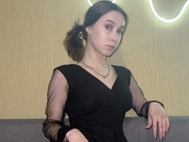 BellaBoom with brown pigtails, wearing a black sheer dress, has small tits, a necklace, and a ring, leaning on a grey couch, with a light-up wall behind.