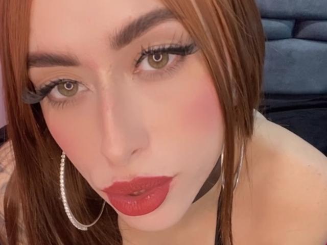 SarahLuxe has red lipstick, thick eyelashes, brown eyes, and brown hair. She's wearing a black choker and silver hoop earrings, with a pink blush on her cheeks.