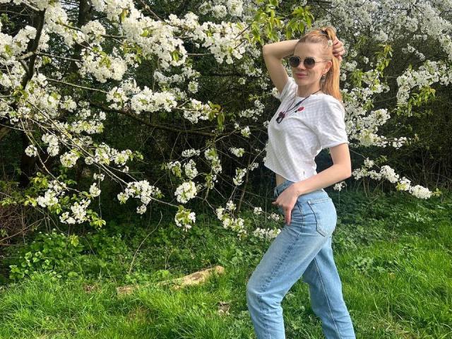 LunaLickies with blonde hair, wearing sunglasses, white tee, and blue jeans, stands in grass, hand in hair, under blooming white flowers, showing her small tits and slim ass.