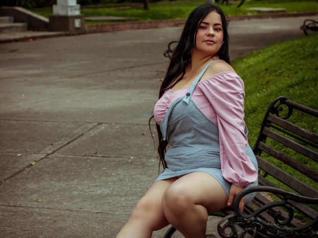 NohayMuller with long black hair, pink off-shoulder top, and gray dress, sitting on a bench, showing her ass, looking confident, outdoors, green grass in the background.