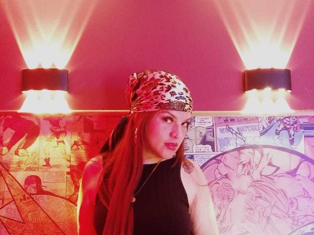 SkylarDesire with long red hair, wearing a leopard print headband, black sleeveless top, and hoop earrings, looking to the side with a slight pout, under pink and yellow lights.