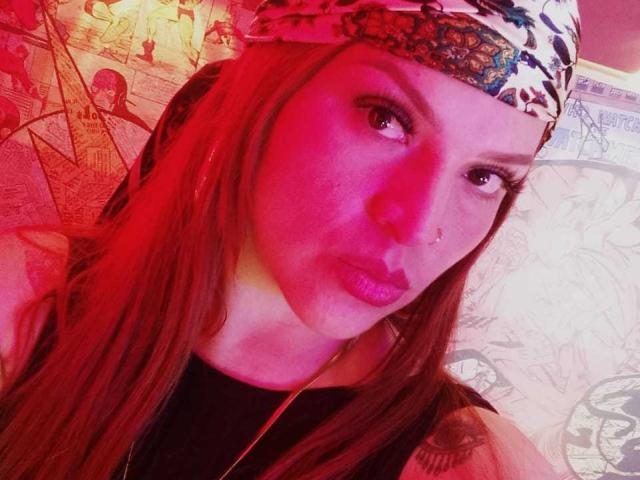 SkylarDesire with red-pink lit face, wearing a floral headscarf, nose ring, and black top, has long brown hair, and glossy pink lips, looking at the camera.