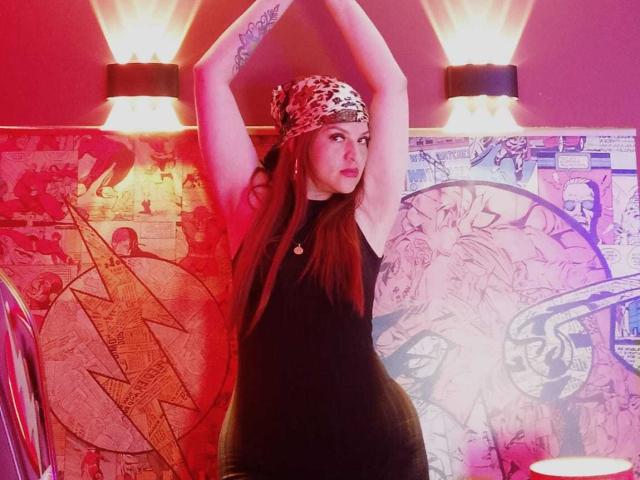 SkylarDesire stands with arms raised, wearing a black dress, leopard print headscarf, and long brown hair, showing tattoos on her arms and a cute face. Red and purple lighting.