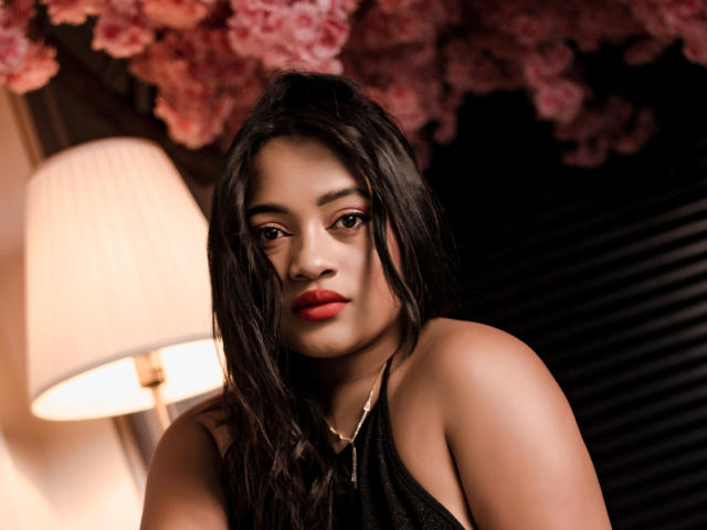 SaritaCute with wet black hair, red lipstick, and black halter top, looking down with serious eyes, standing next to a lit lamp and pink cherry blossoms.