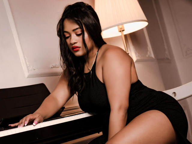SaritaCute, with curvy ass, large tits, and dark hair, sits at a piano, wearing a tight black dress, looking down with red lipstick, under a warm lamp.