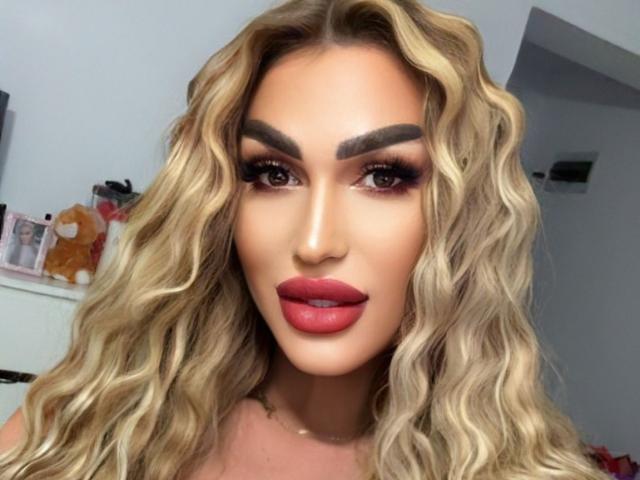 NicolSNaya has blonde wavy hair, thick eyebrows, dark red lipstick, and smoky eye makeup, with a serious expression. Her tits are not visible in this image.