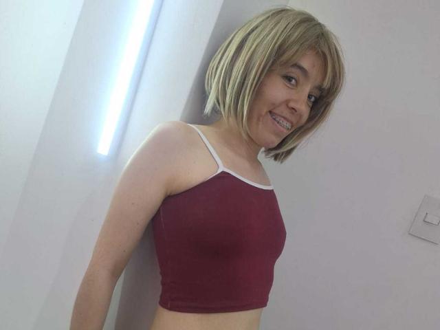 ElodiePerrine with short blonde hair, smiling, wearing a red crop top with white trim, exposing her small tits, standing against a white wall.