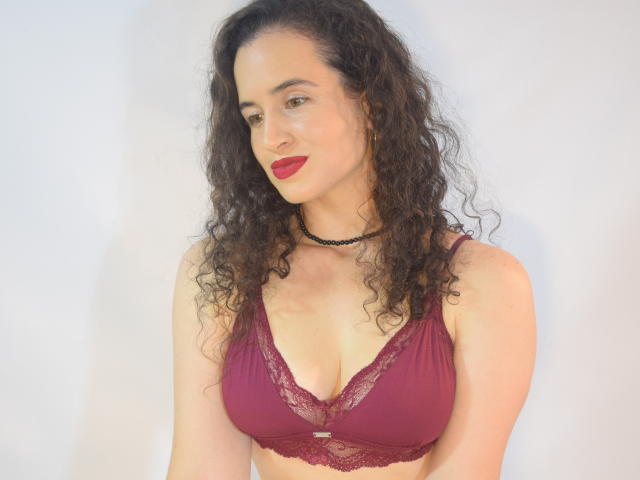 BrenditaSecret with curly dark hair, red lipstick, and maroon lace bra, showing her tits, wearing a black choker, looking to the side, smiling slightly.