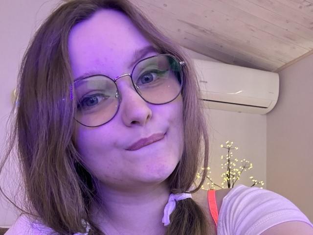 MagikPeachh has pale skin, brown hair, glasses, and a small smile. She's wearing a white shirt with pink straps and a hair tie on her right side.