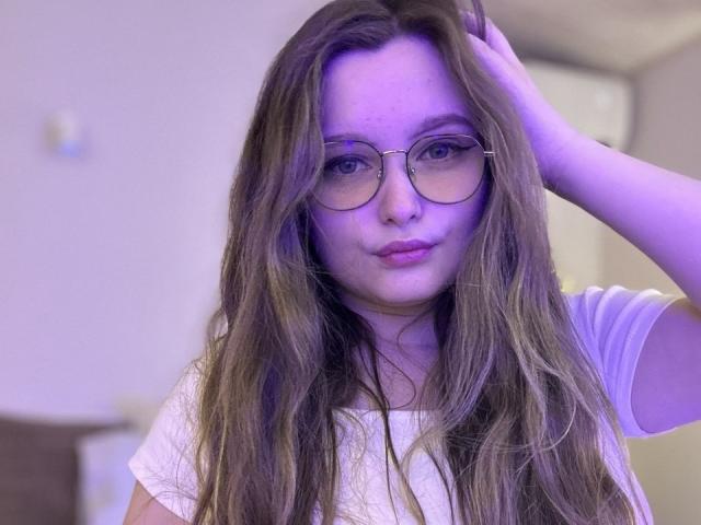 MagikPeachh with long brown hair, glasses, and a white shirt, touching her head with a purple glow on her face. She's looking at the camera with a slight smile.