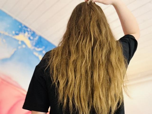 MagikPeachh with long, wavy brown hair, wearing a black shirt, raising her right arm, showing her back. Her hand is in her hair, ceiling visible.