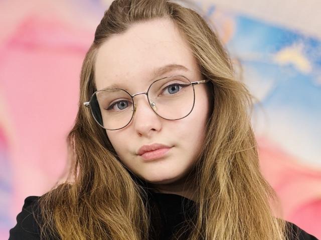 MagikPeachh has pale skin, blue eyes, and wavy blonde hair. She's wearing round glasses and a black top, with a neutral expression and pouty lips.