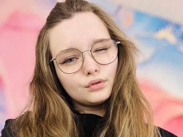MagikPeachh, avec un œil qui cligne, des lunettes rondes et des cheveux bruns clairs, portant un haut noir, a un visage légèrement boudeur, pâle, avec des taches de rousseur et un maquillage subtil..