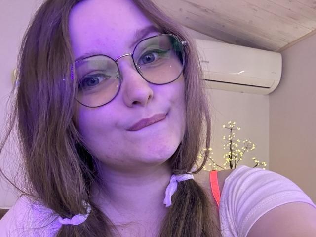 MagikPeachh tem a pele com tons de roxo, cabelo castanho em maria-chiquinhas com fitas rosa, usa óculos e uma camisa branca, sorrindo com as bochechas levemente coradas. Ar condicionado ao fundo..