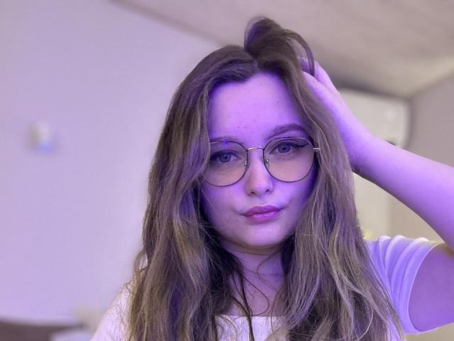 MagikPeachh, avec de longs cheveux bruns, portant des lunettes et une chemise blanche, a une main dans ses cheveux, avec un éclairage violet et un léger froncement de sourcils, regardant directement l'appareil photo..
