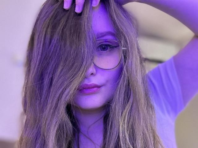 MagikPeachh with long, wavy brown hair covering one eye, wearing glasses, purple lipstick, and a white shirt, under blue lighting, hand in hair, looking at camera.