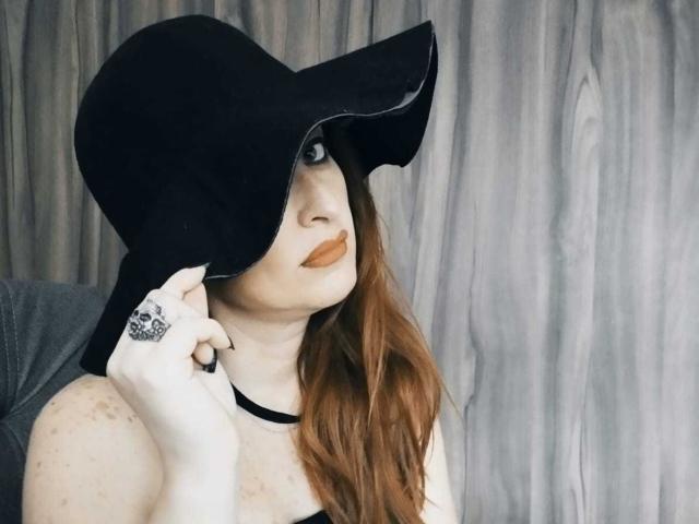 NoctisEye with red hair, wearing a black wide-brim hat, black choker, and skull ring, looking up with orange lipstick, holding hat, showing freckled arms.