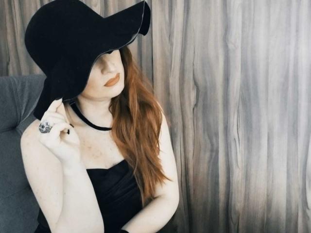 NoctisEye with pale skin, red hair, wearing a black hat, strapless black dress, choker, and ring, touching hat, with orange lipstick, sitting on a gray chair.