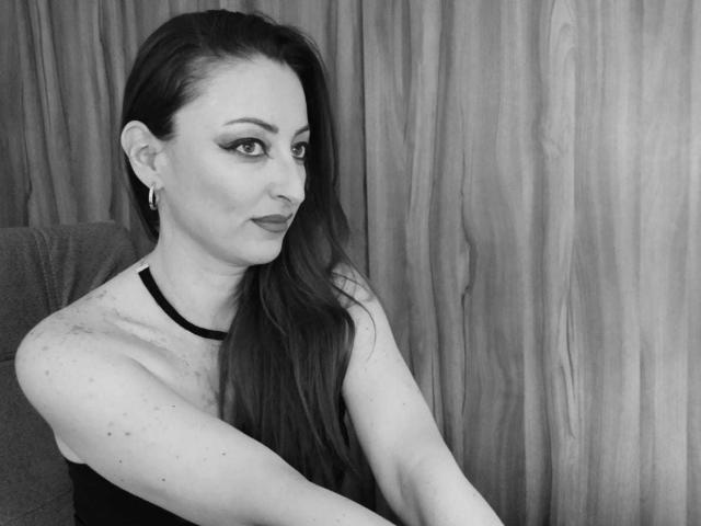 NoctisEye has long dark hair, wearing a black sleeveless top, showing freckles on her arm, with eyeliner and dark lipstick, sitting in a chair, looking to the side.