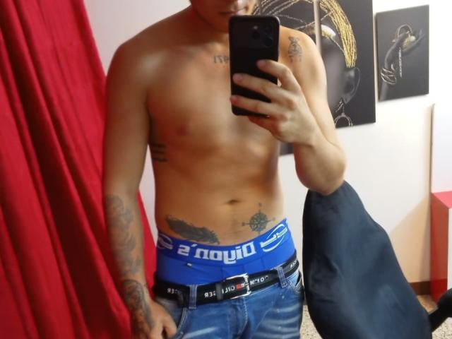 ThommyHills, shirtless with tattoos, wearing blue Reebok underwear, holding a phone, jeans with black belt, red curtain, black chair, and wall art in background.