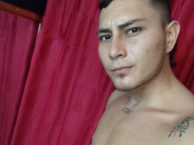 ThommyHills, shirtless with a short haircut, has a tattoo on his right shoulder, a number tattoo on his left chest, and a serious expression, standing against red curtains.
