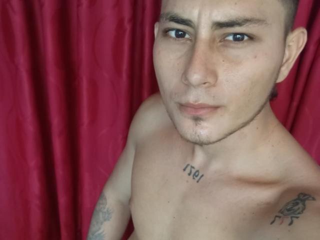 ThommyHills, shirtless, with short brown hair, light skin, and tattoos on chest and shoulder, has a neutral expression, blue eyes, and a small goatee, against a red curtain.