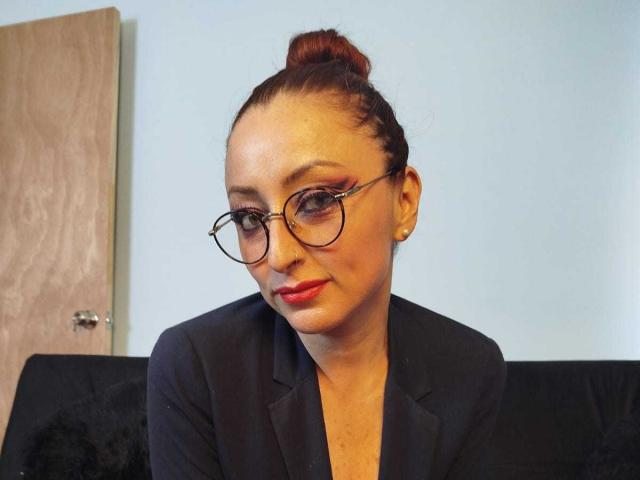 NoctisEye with red hair in a bun, wearing glasses, black blazer, and red lipstick, sitting on a black couch, looking at camera with a slight smile.