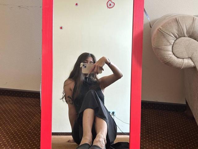 Mirror selfie of MoonLiza with long brown hair, black dress, tattoo on right arm, holding camera to face, legs crossed, sitting on carpet, red mirror frame, beige couch.