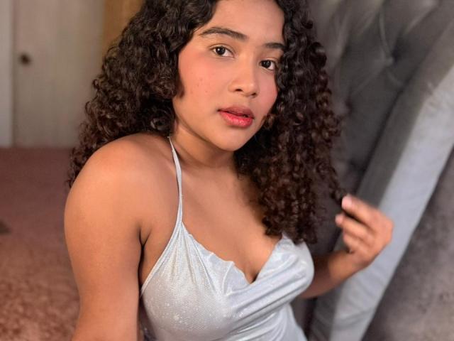 AnnyRestrepo with curly brown hair, wearing a shiny silver dress, has a cute face, red lips, and small tits, sitting on a grey tufted chair.