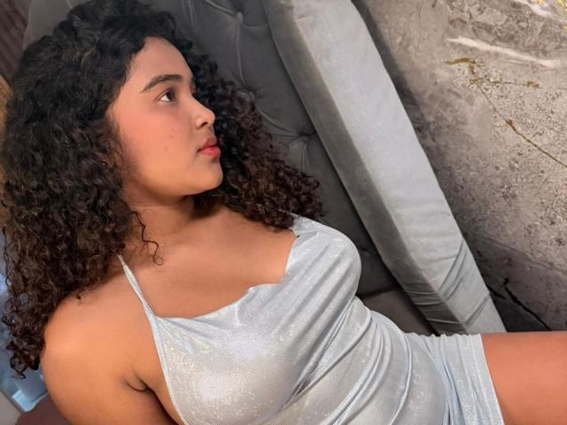 AnnyRestrepo with curly black hair, wearing a shiny silver halter dress, sitting on a grey tufted chair, gazing to the side, showing her small tits and flat ass.