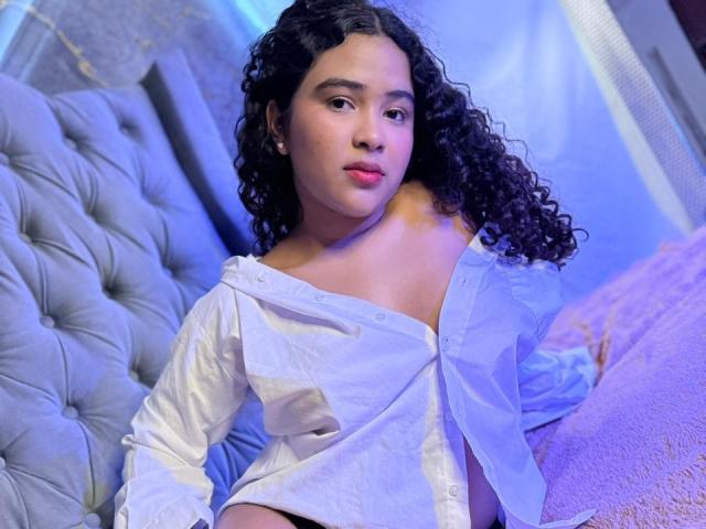AnnyRestrepo with curly black hair, wearing an off-shoulder white shirt, sitting on a tufted grey bed, blue and purple lighting, looking slightly to the side, pink lips.