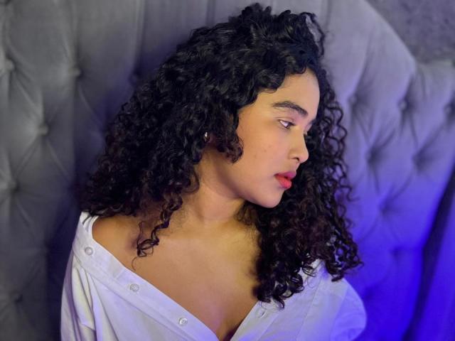 AnnyRestrepo with curly black hair, tan skin, and red lipstick, wearing a white shirt with buttons, looking to the side, against a tufted gray background.