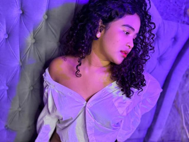 AnnyRestrepo with curly dark hair, off-shoulder white shirt, and pinkish-blue light, looking to the side, showing her tits and small earrings, sitting on a tufted, light-gray sofa.