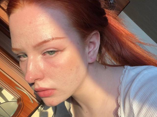 CindyNeonx has pale skin, red hair in a ponytail, green eyes, and a slightly freckled face with visible pores and a small nose. She's wearing a white top.