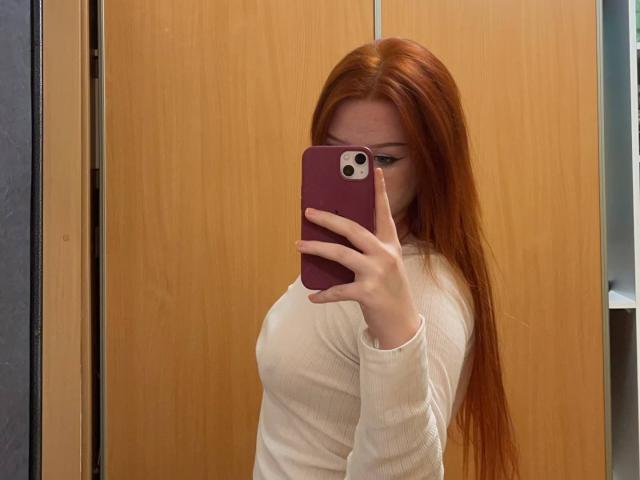 CindyNeonx with long red hair, white ribbed shirt, holding purple phone, taking mirror selfie, showing her tits. Wooden door background, visible from side.