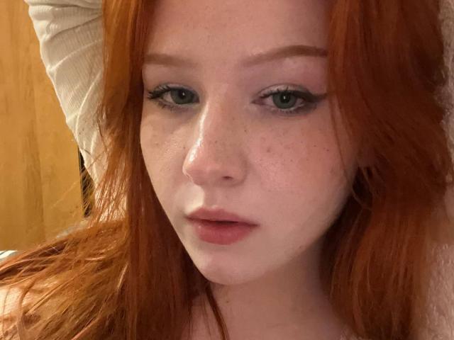 CindyNeonx has red hair, green eyes, and freckles on her fair skin. She's looking at the camera with a slight pout, wearing a white long-sleeve shirt.