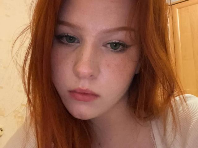 CindyNeonx has red hair, green eyes, and freckles with slightly parted pink lips. She's wearing a white top, looking straight at the camera with a neutral expression.
